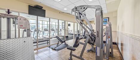 A gym with various exercise equipment, including weight machines and treadmills, with large windows allowing natural light.