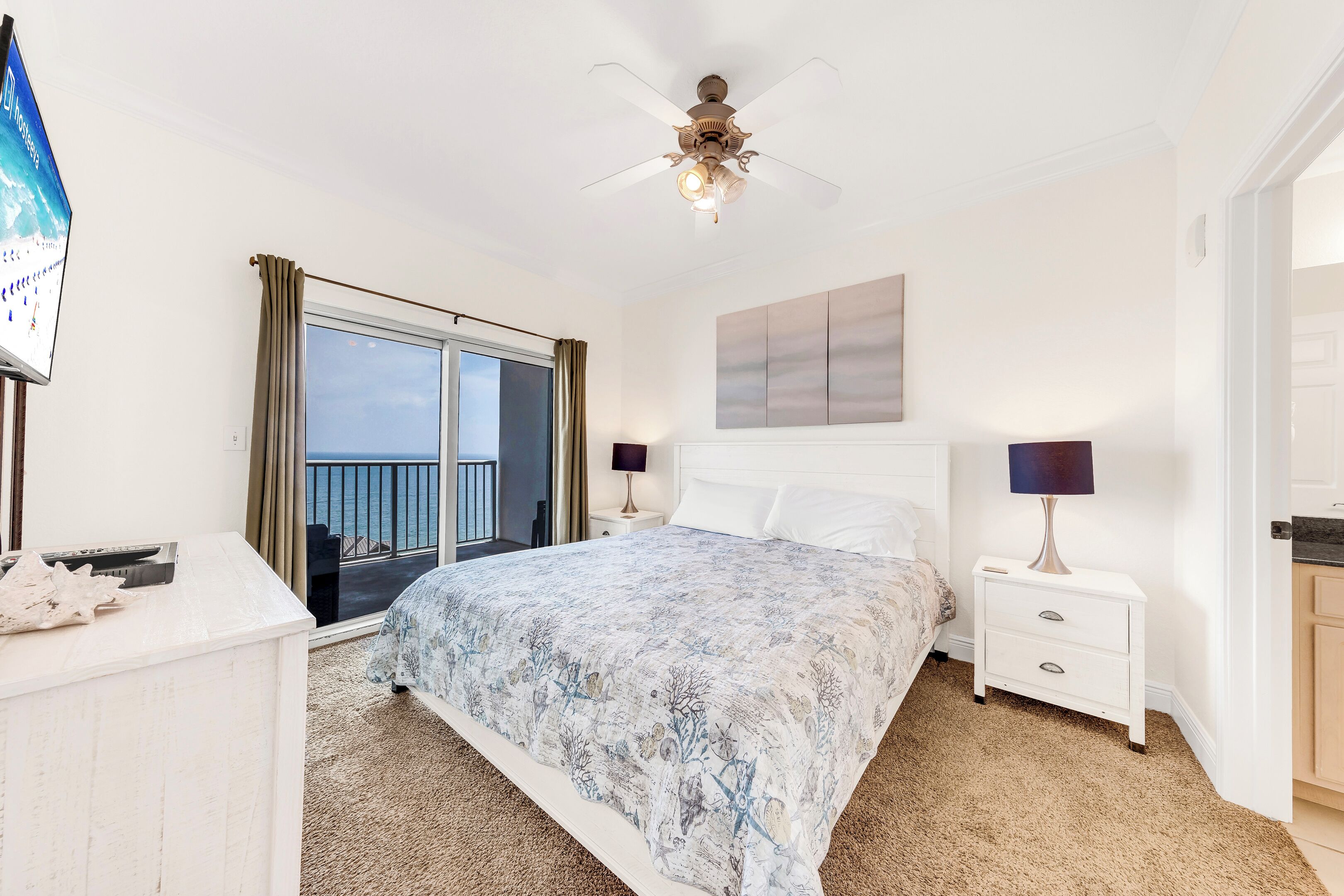 A bedroom with a bed, two bedside tables, a ceiling fan, a large window with curtains, and a view of the sea.