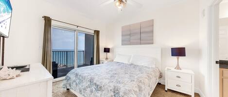 A bedroom with a bed, two bedside tables, a ceiling fan, a large window with curtains, and a view of the sea.