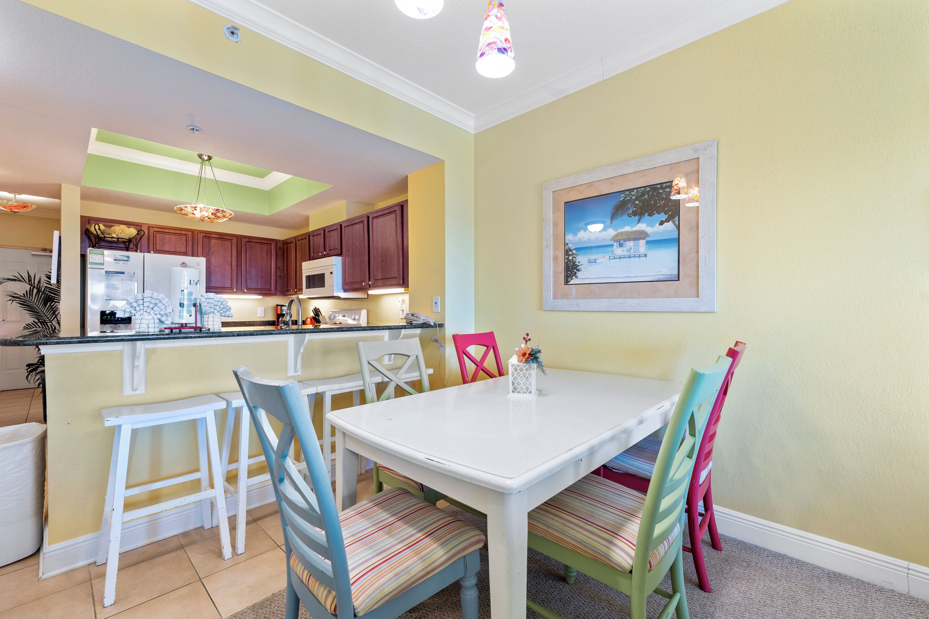Un comedor con una mesa blanca y sillas coloridas, una cocina al fondo y un cuadro en la pared.