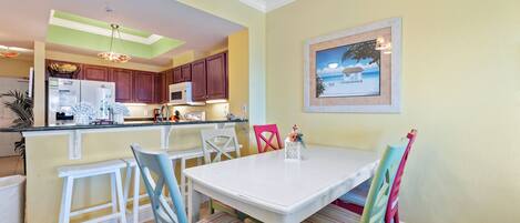 Un comedor con una mesa blanca y sillas coloridas, una cocina al fondo y un cuadro en la pared.