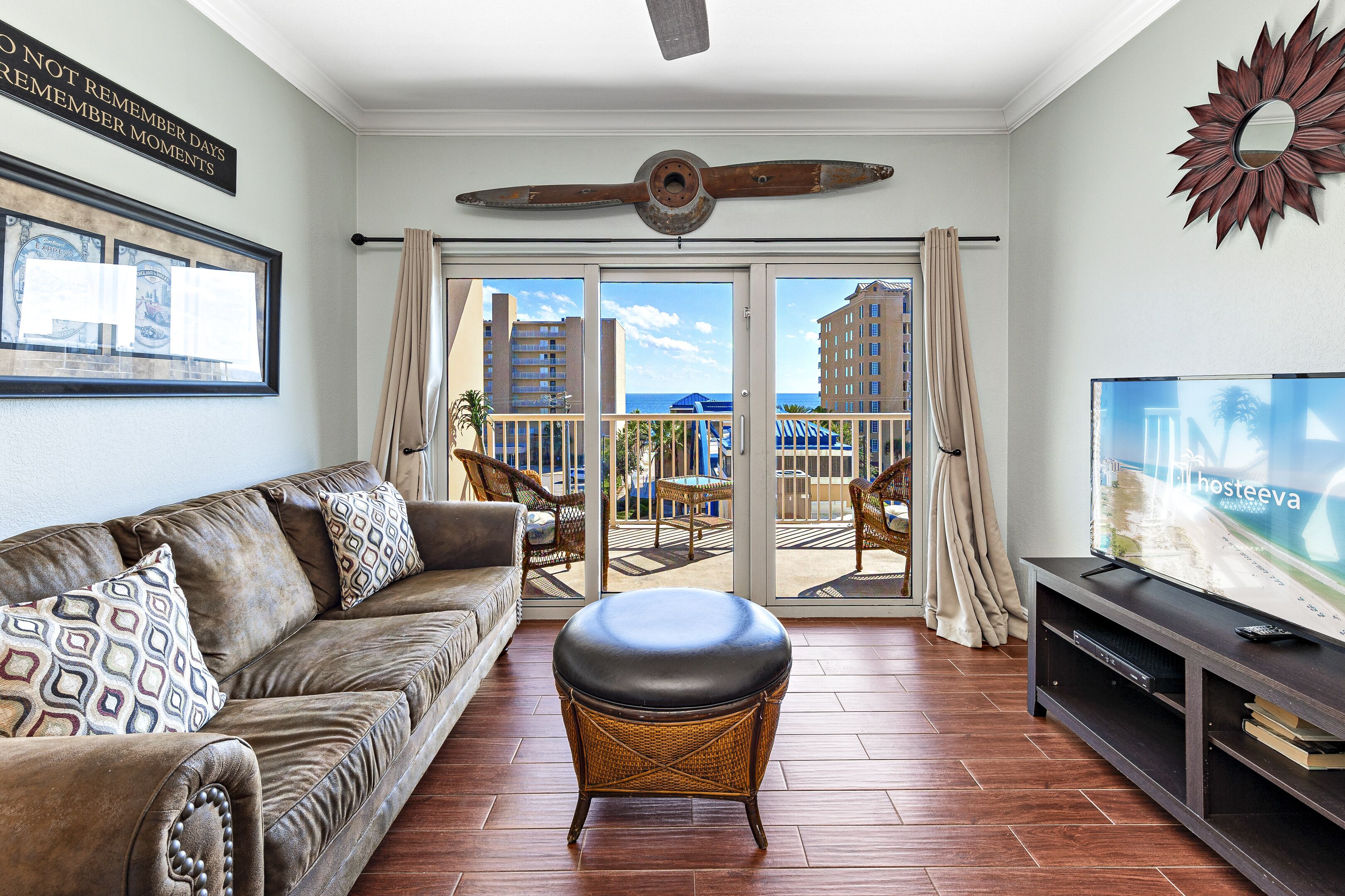 A living room with a leather sofa, a wicker ottoman, a flat-screen TV, and a balcony with a view of the city and ocean.