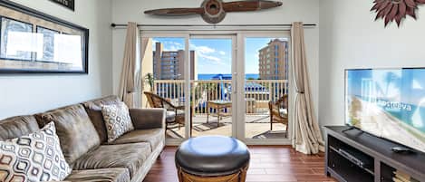A living room with a leather sofa, a wicker ottoman, a flat-screen TV, and a balcony with a view of the city and ocean.