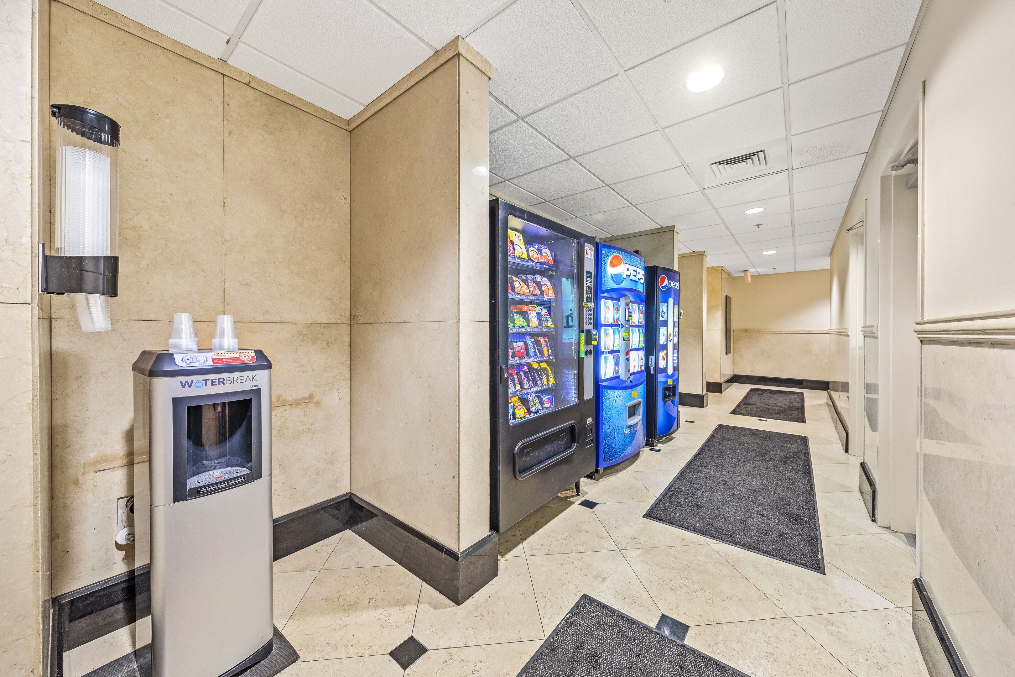 A hotel corridor with vending machines, including a water dispenser and a Pepsi machine, and a tiled floor with black and beige tiles.