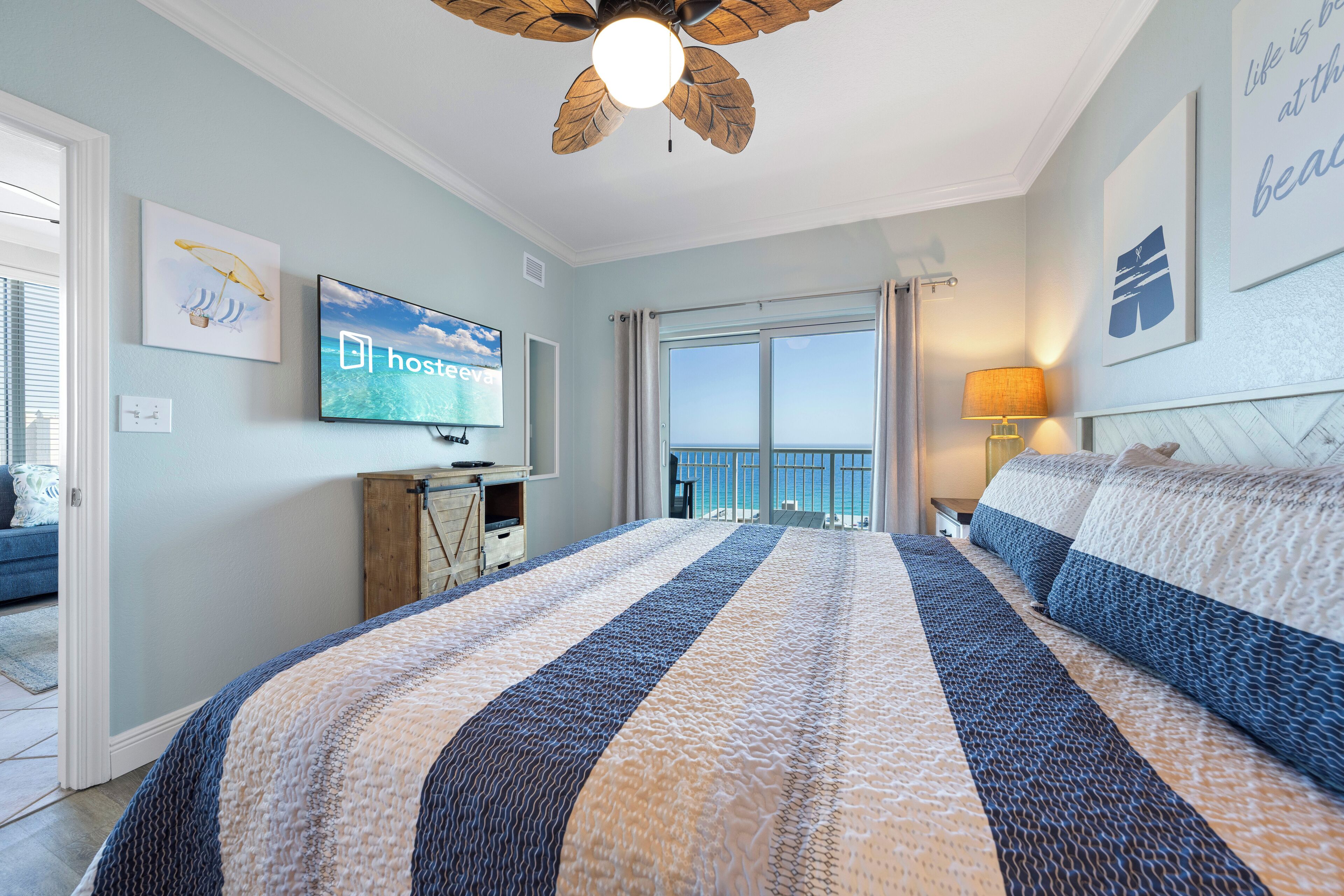 Habitación de hotel con una cama grande, ventilador de techo, televisor en la pared y vista al mar por la ventana.