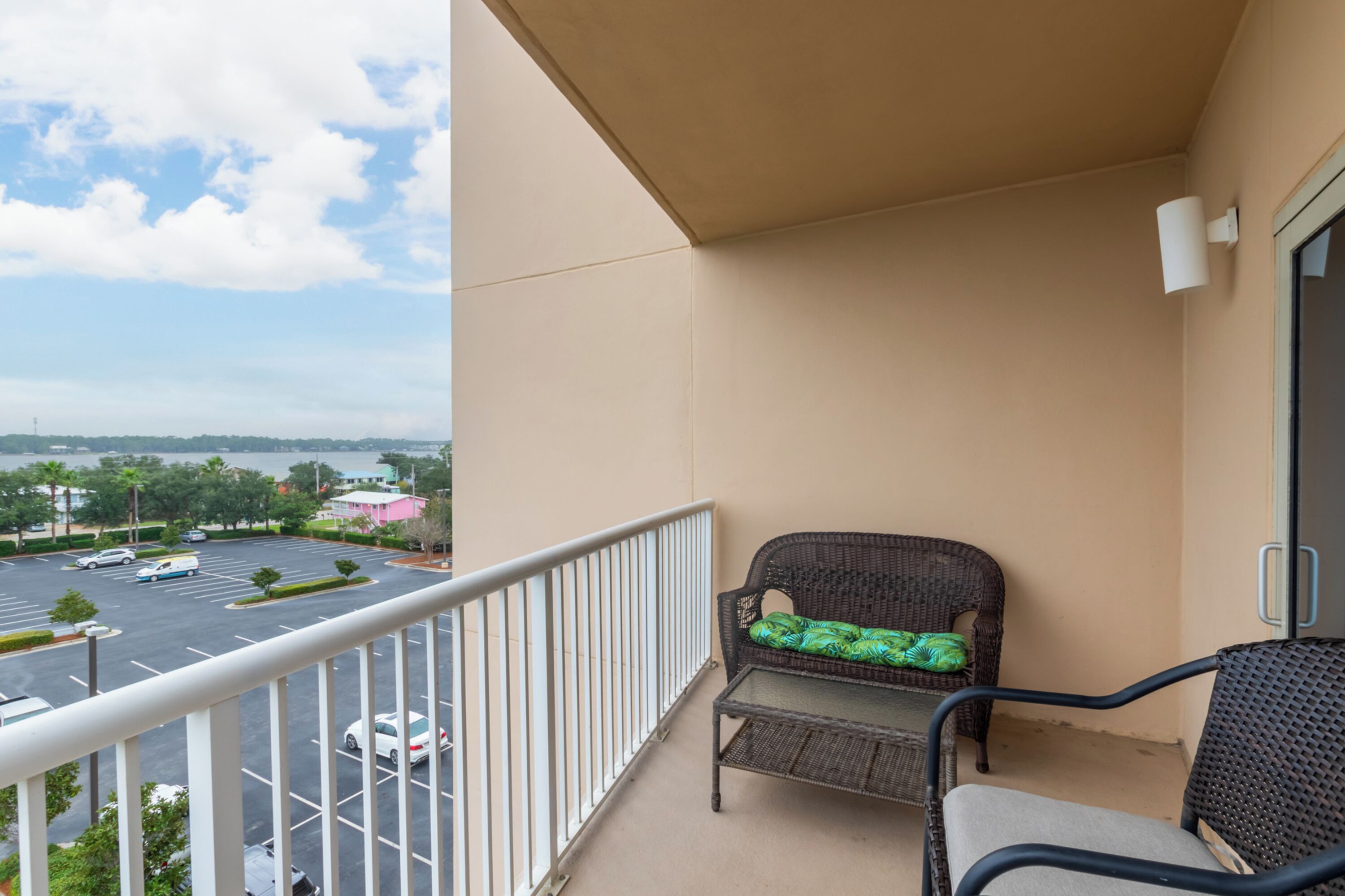 A balcony with a wicker chair and a table, overlooking a parking lot and distant buildings.