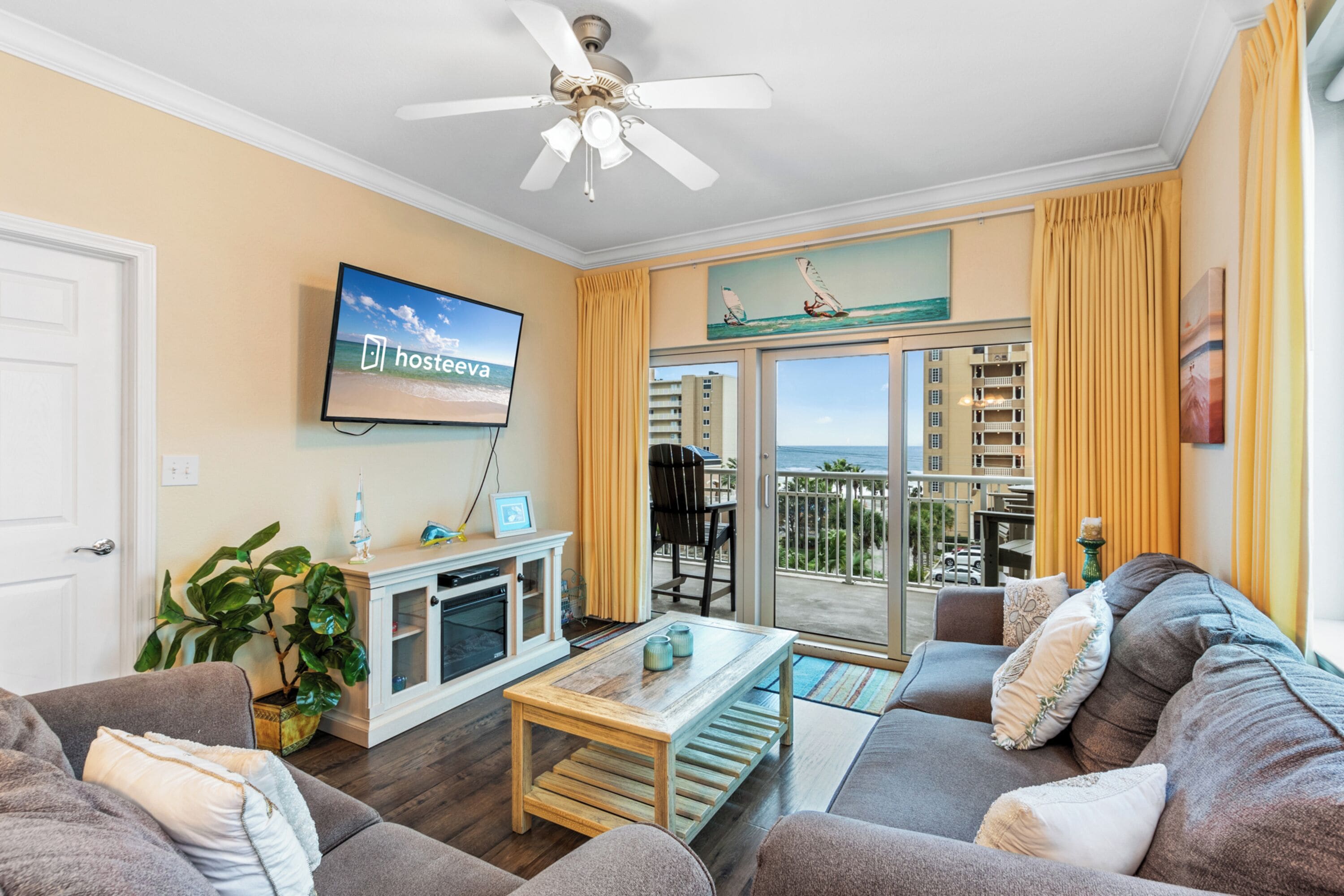 A living room with a television, a ceiling fan, a sofa, a coffee table, and a view of the ocean through a sliding glass door.