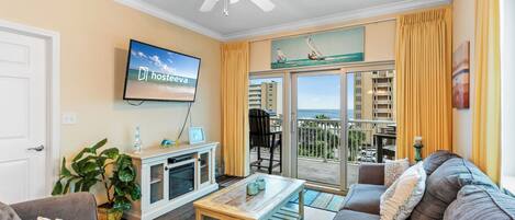 A living room with a television, a ceiling fan, a sofa, a coffee table, and a view of the ocean through a sliding glass door.