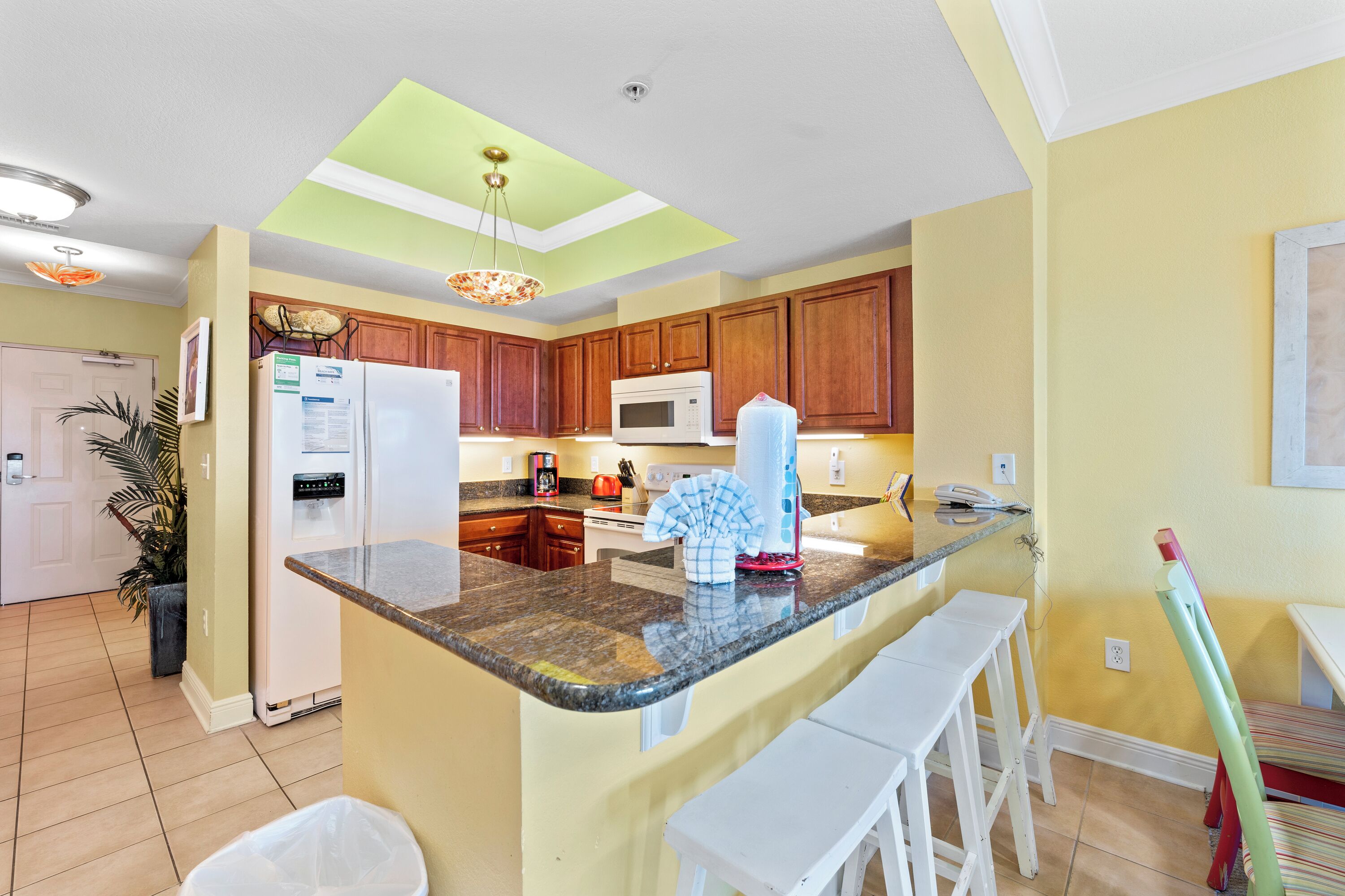 A kitchen with wooden cabinets, a granite countertop, a white refrigerator, a microwave, and a hanging light fixture.
