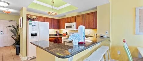 A kitchen with wooden cabinets, a granite countertop, a white refrigerator, a microwave, and a hanging light fixture.