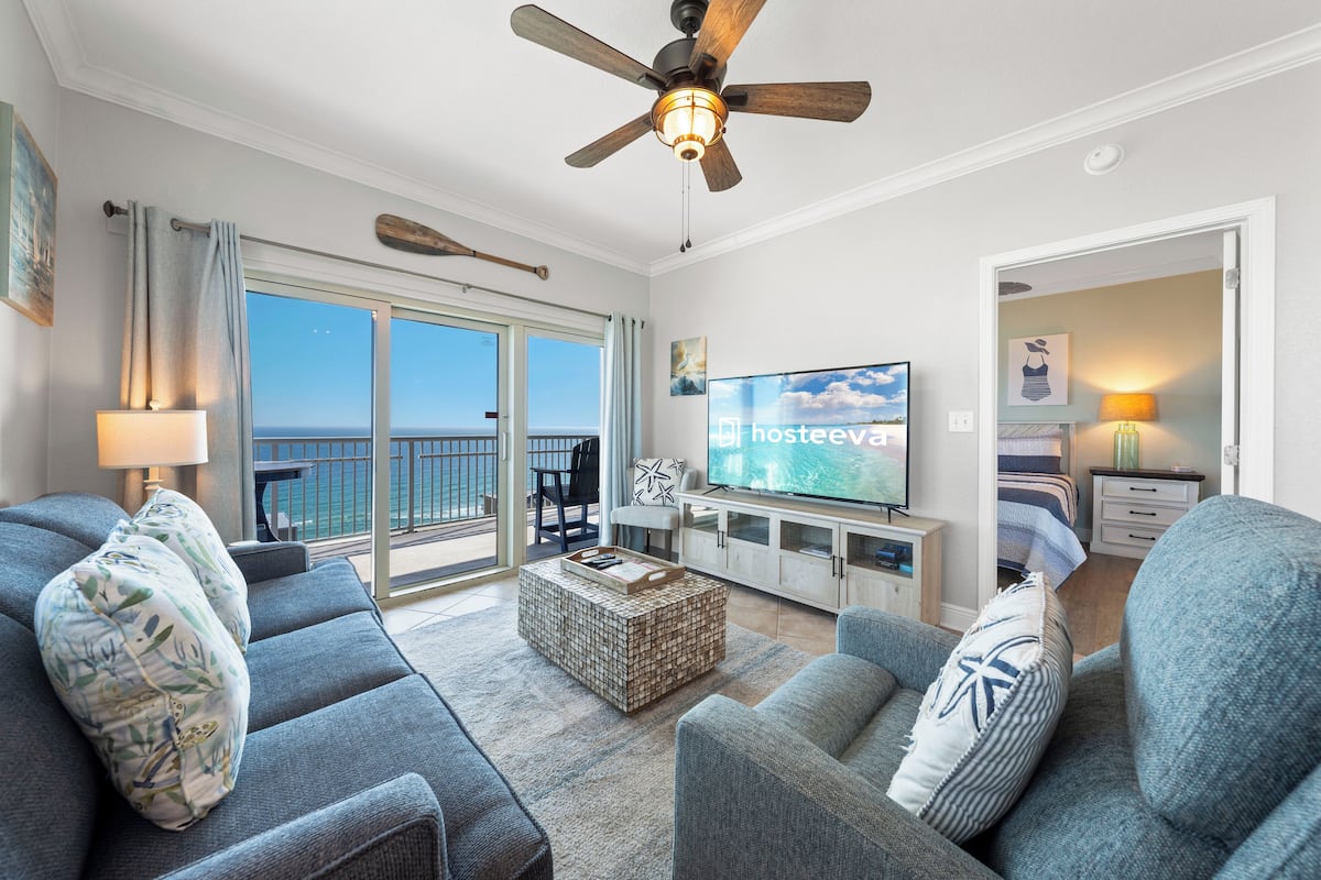 A modern living room with a large TV, comfortable seating, a ceiling fan, and a balcony with a view.