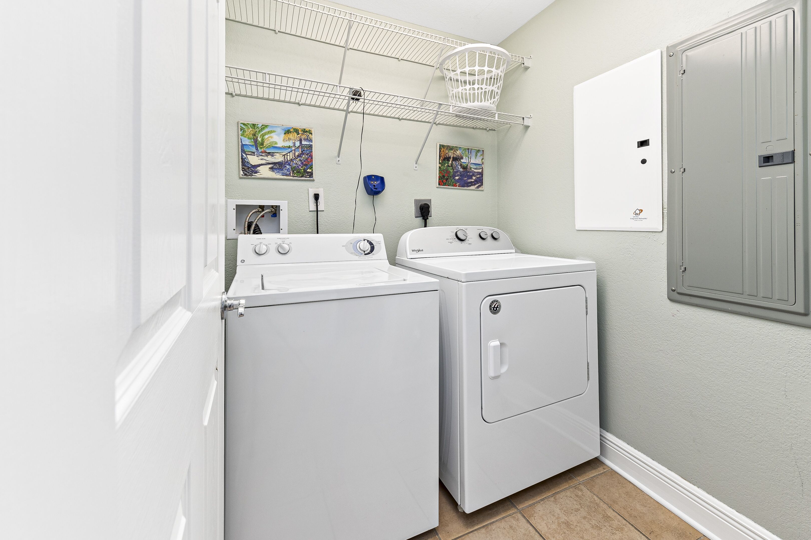 A laundry room with two white washers and dryers, a metal shelf, and two wall-mounted paintings.