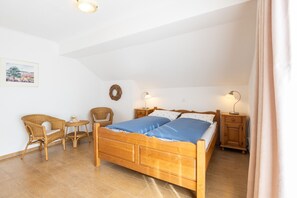 Une chambre à coucher avec un lit en bois, deux tables de chevet, des chaises en osier, une petite table ronde et un luminaire fixé au mur.
