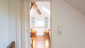A hallway leading to a room with a sloped ceiling, wooden flooring, and a window with striped curtains.