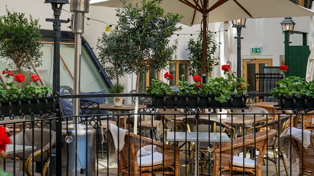 Zona de café al aire libre con mesas y sillas, rodeada por una verja metálica y decorada con plantas en macetas y una farola.
