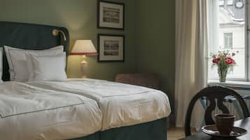 Una cama bien hecha con sábanas blancas, una lámpara de noche, dos cuadros en la pared, una ventana con vistas a edificios y una mesa con una taza y un plato.