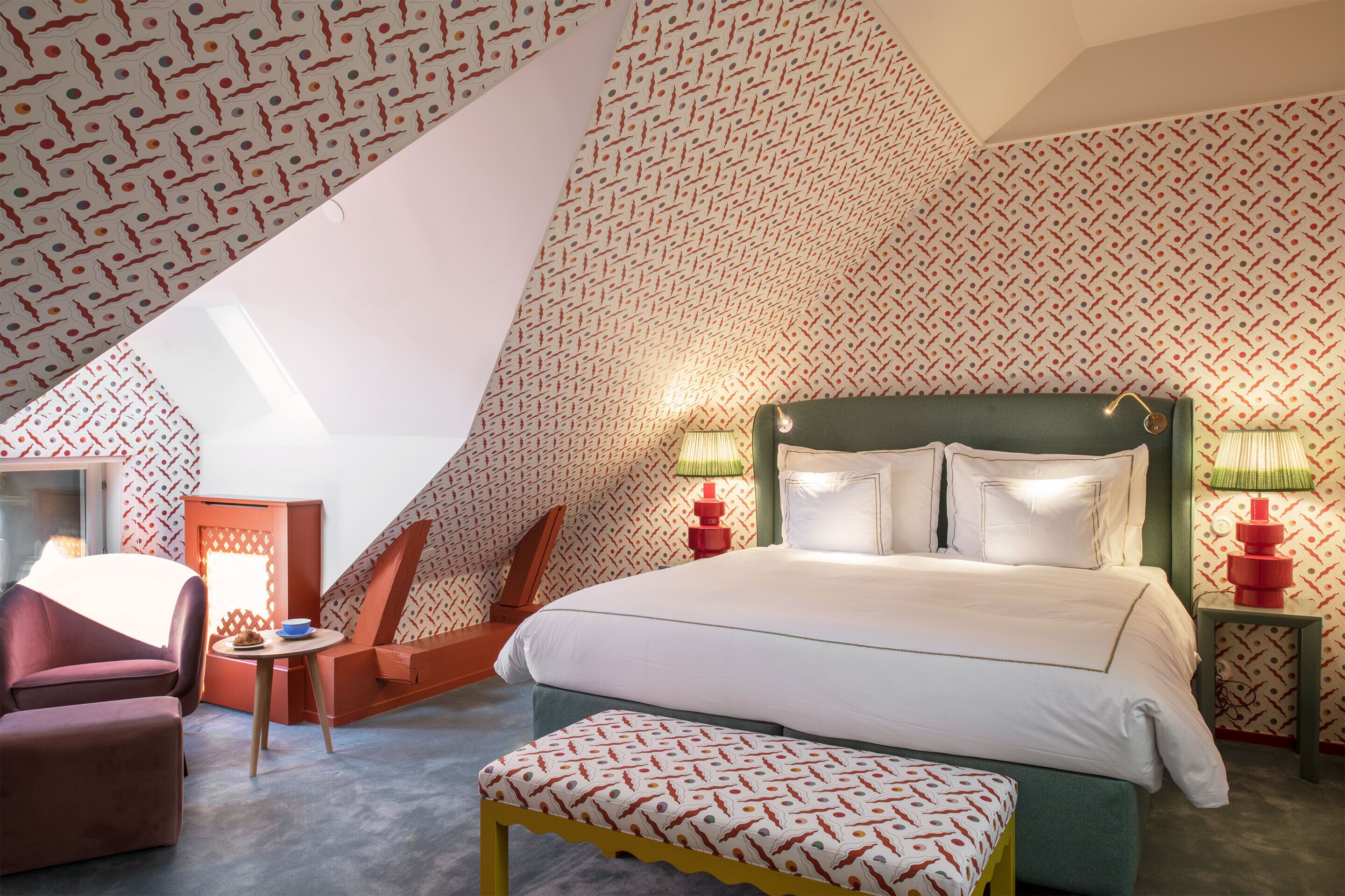 A bedroom with a sloped ceiling, patterned wallpaper, a bed with white linens, two bedside lamps, a green headboard, and a small table with a bowl.