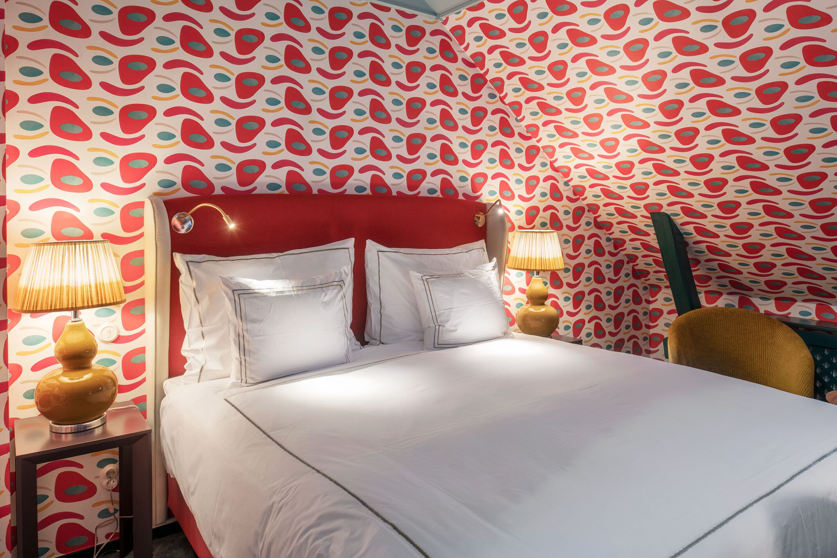 Quarto de hotel com cabeceira vermelha, roupa de cama branca e papel de parede estampado. Há duas luminárias de mesa e uma cadeira de madeira.