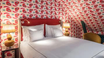 Quarto de hotel com cabeceira vermelha, roupa de cama branca e papel de parede estampado. Há duas luminárias de mesa e uma cadeira de madeira.
