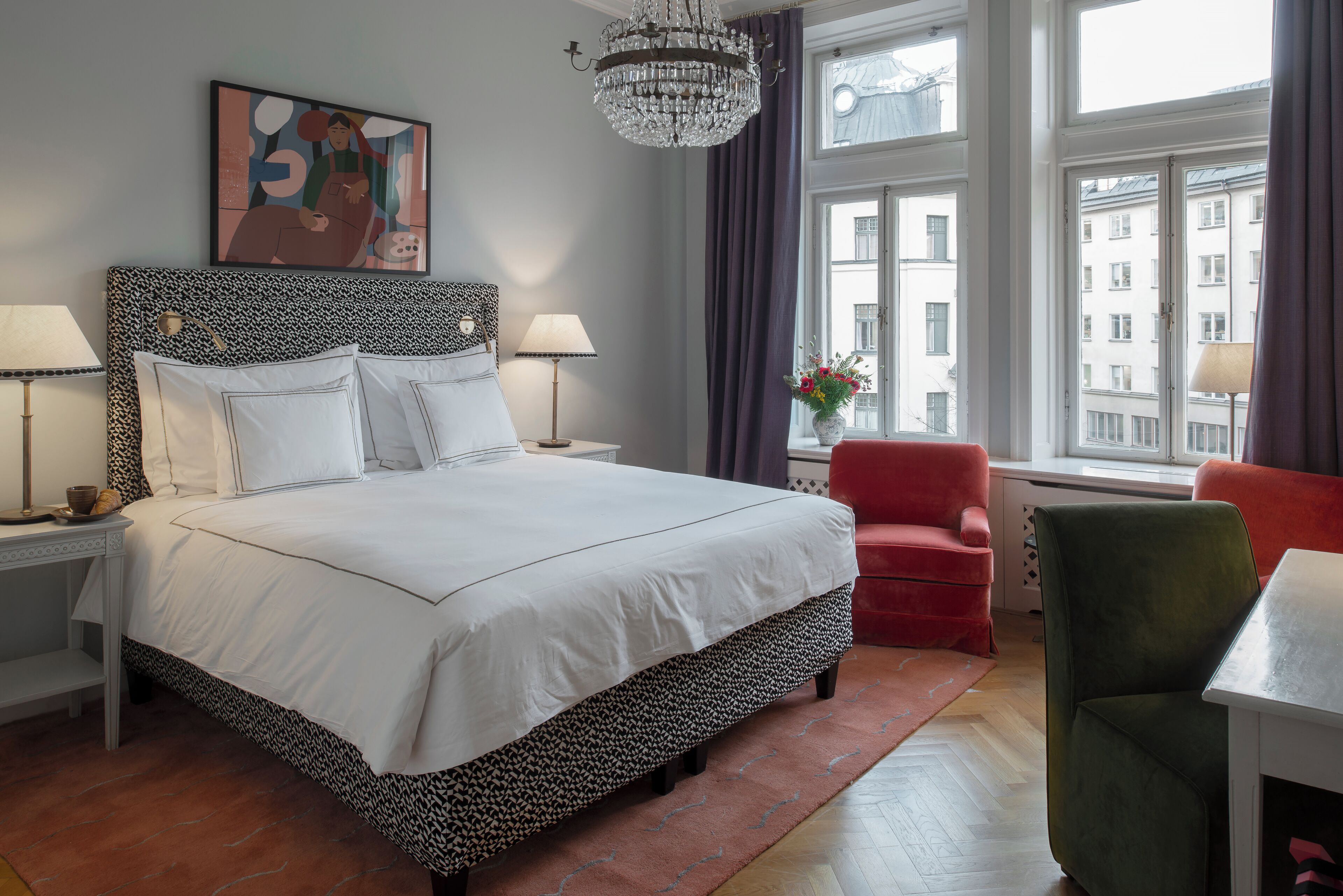 Una habitación de hotel con una cama grande, dos lámparas de noche, un candelabro, una silla roja, una silla verde y un ventanal con vistas a la ciudad.
