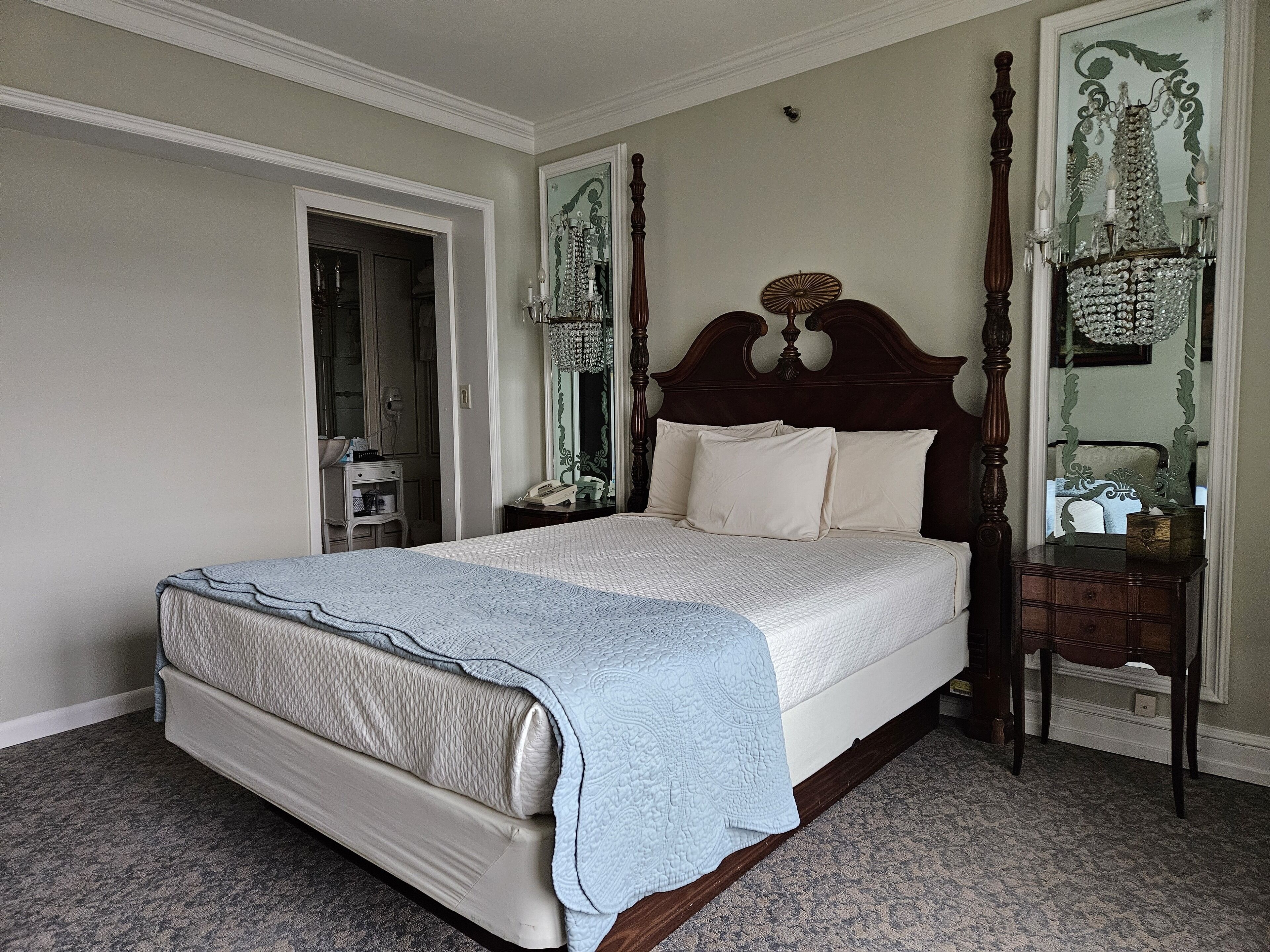 Una cama con dosel y tablero de madera, una mesita de noche con espejo y otra con lámpara.