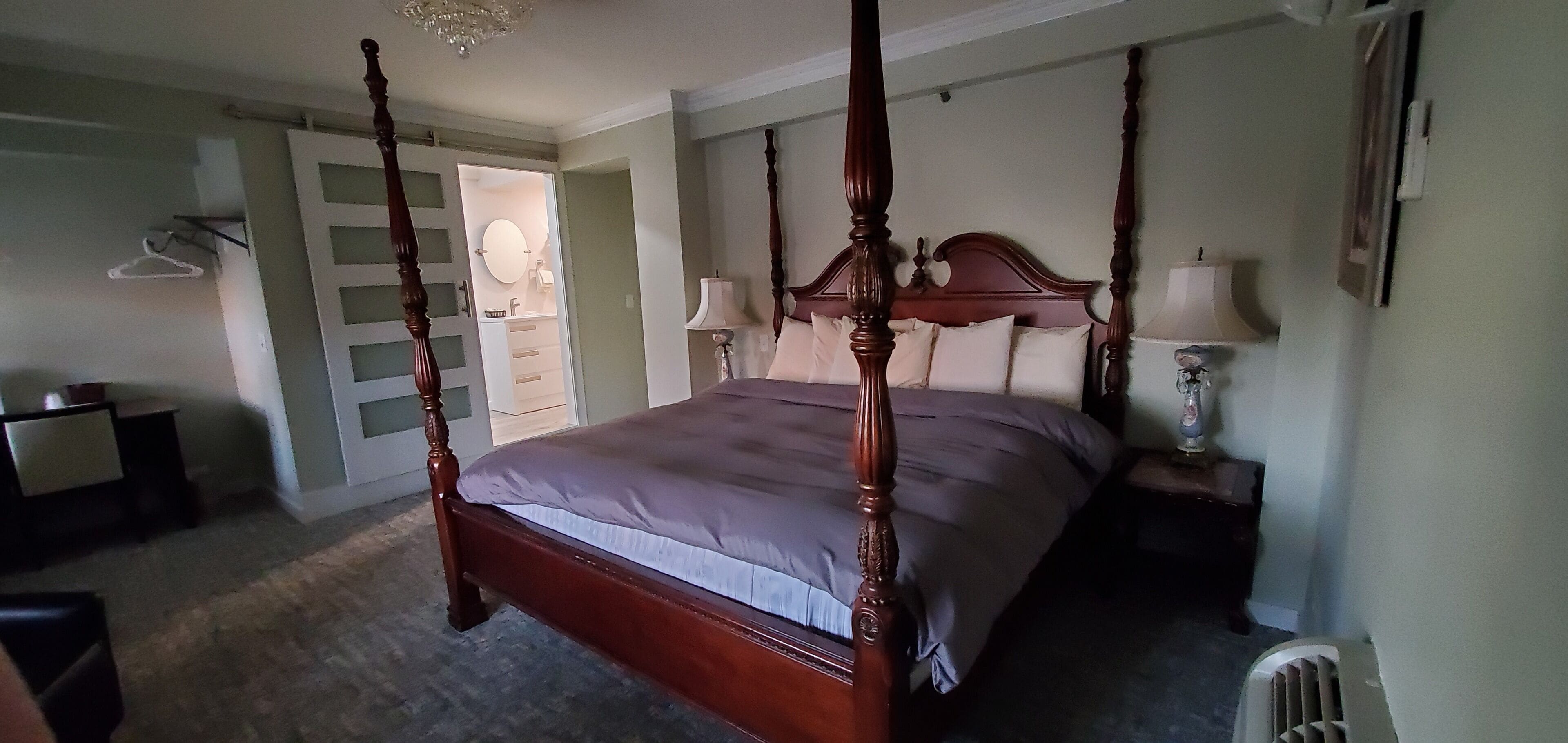Una cama con dosel y estructura de madera, una mesita de noche con lámpara, y una puerta que da a un baño.