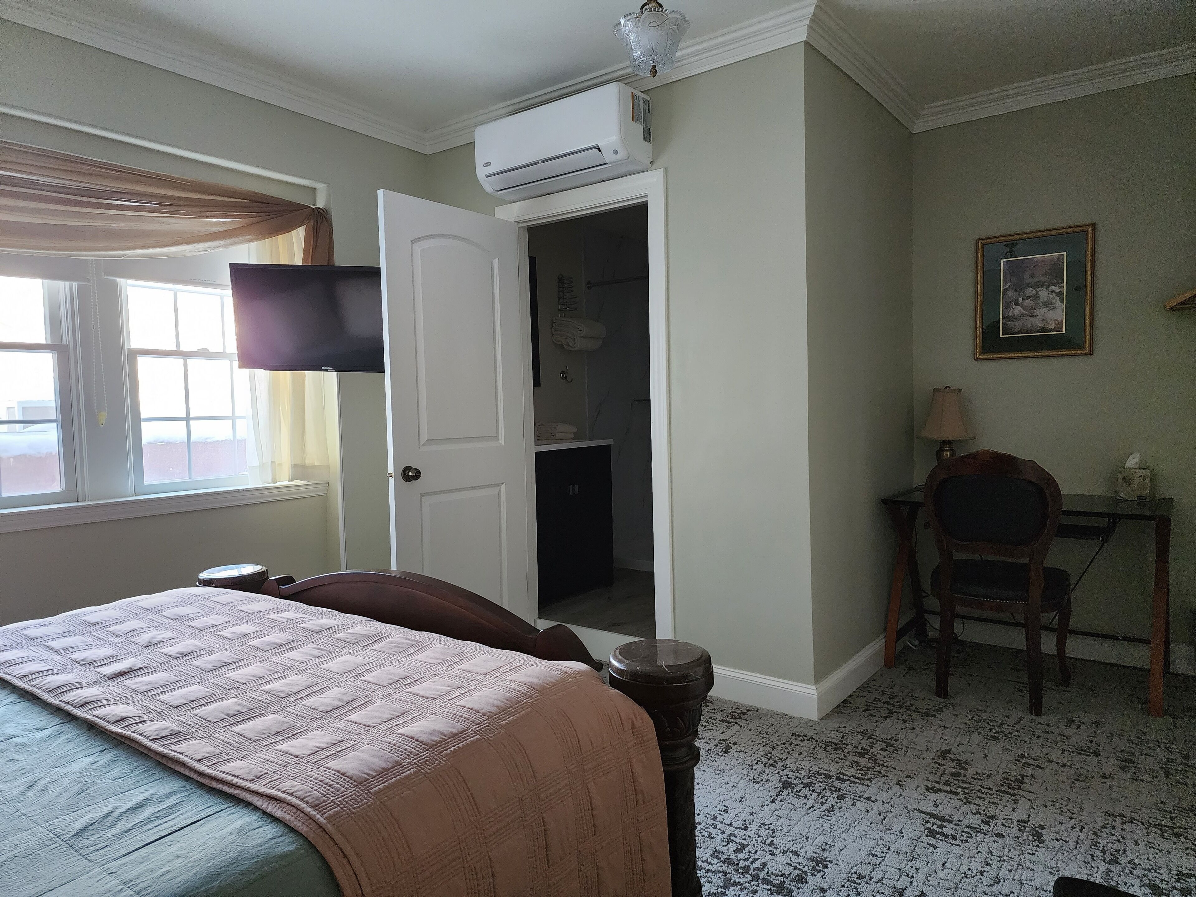 Una habitación de hotel con cama, escritorio, silla, equipo de aire acondicionado, un cuadro enmarcado y una ventana con cortinas.