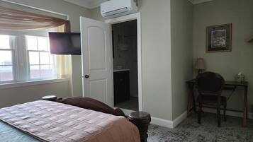 Una habitación de hotel con cama, escritorio, silla, equipo de aire acondicionado, un cuadro enmarcado y una ventana con cortinas.