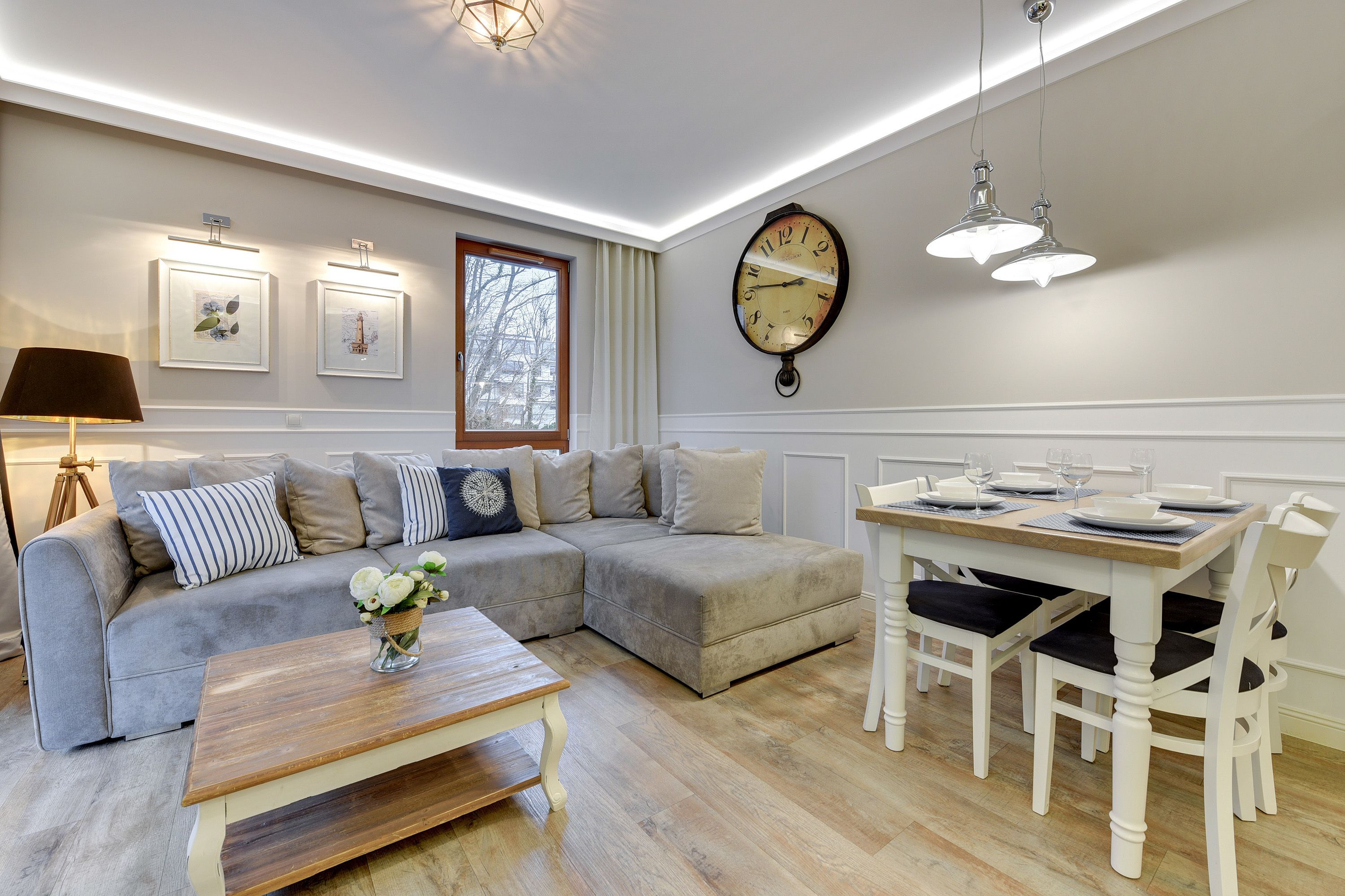 A modern living room with a sectional sofa, a dining table with chairs, a wooden coffee table, wall-mounted artwork, and a large wall clock.