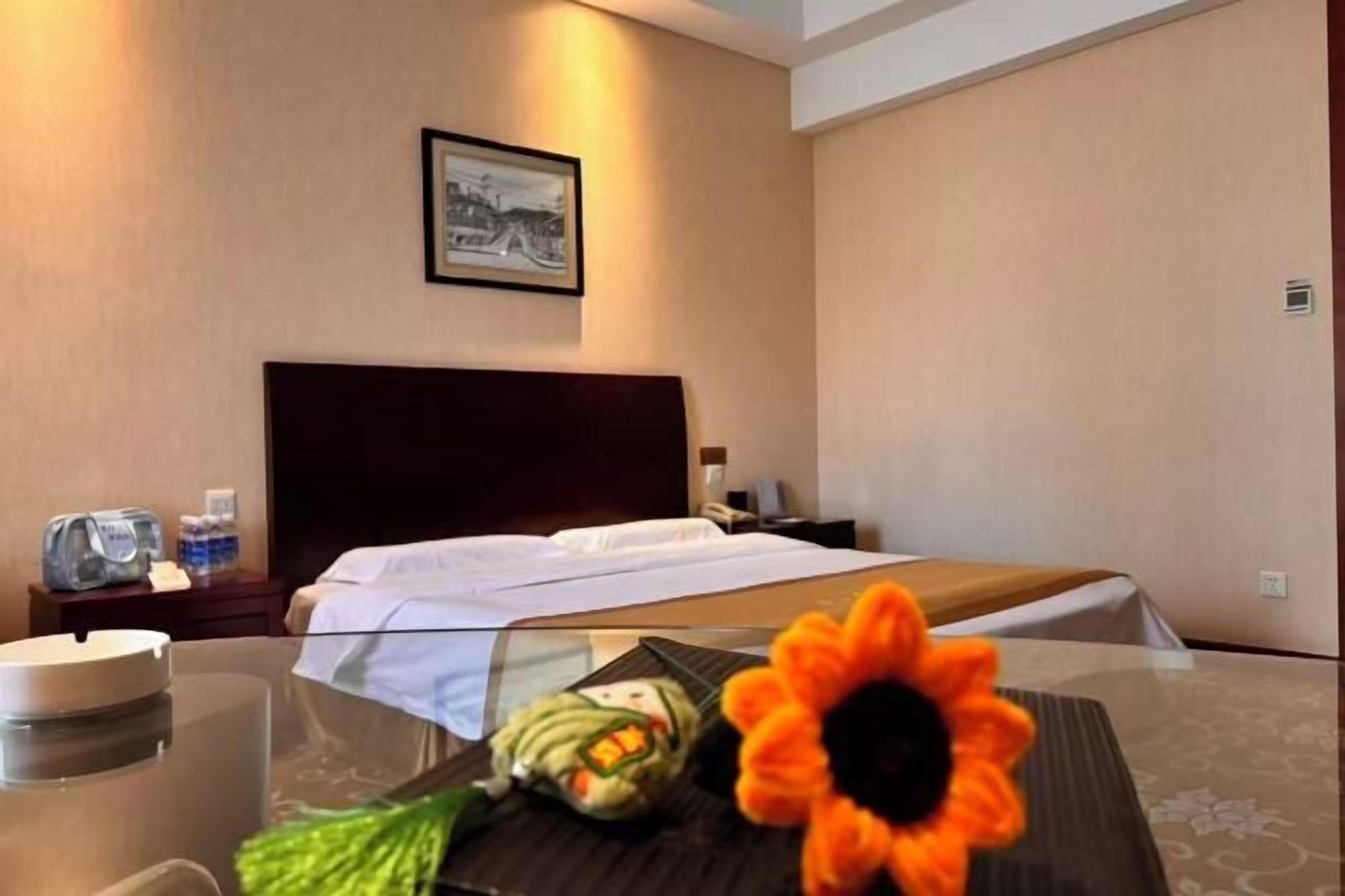 A hotel room with a bed, a nightstand, a framed picture on the wall, and a table with a flower and a bottle.