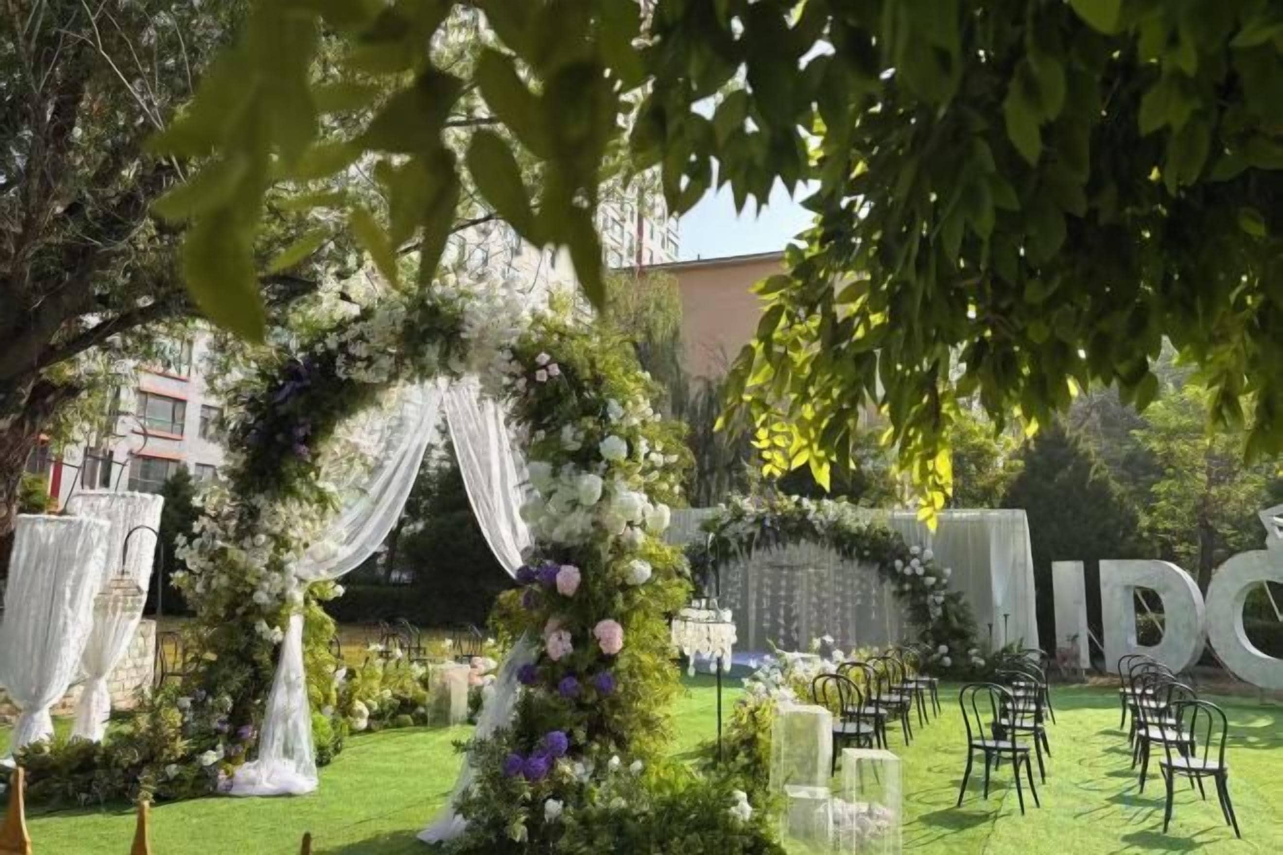 Un lieu de réception en plein air pour un mariage, agrémenté d’une arche fleurie, d’un aménagement pour les invités et d’un décor où figure le mot « MARIAGE ».