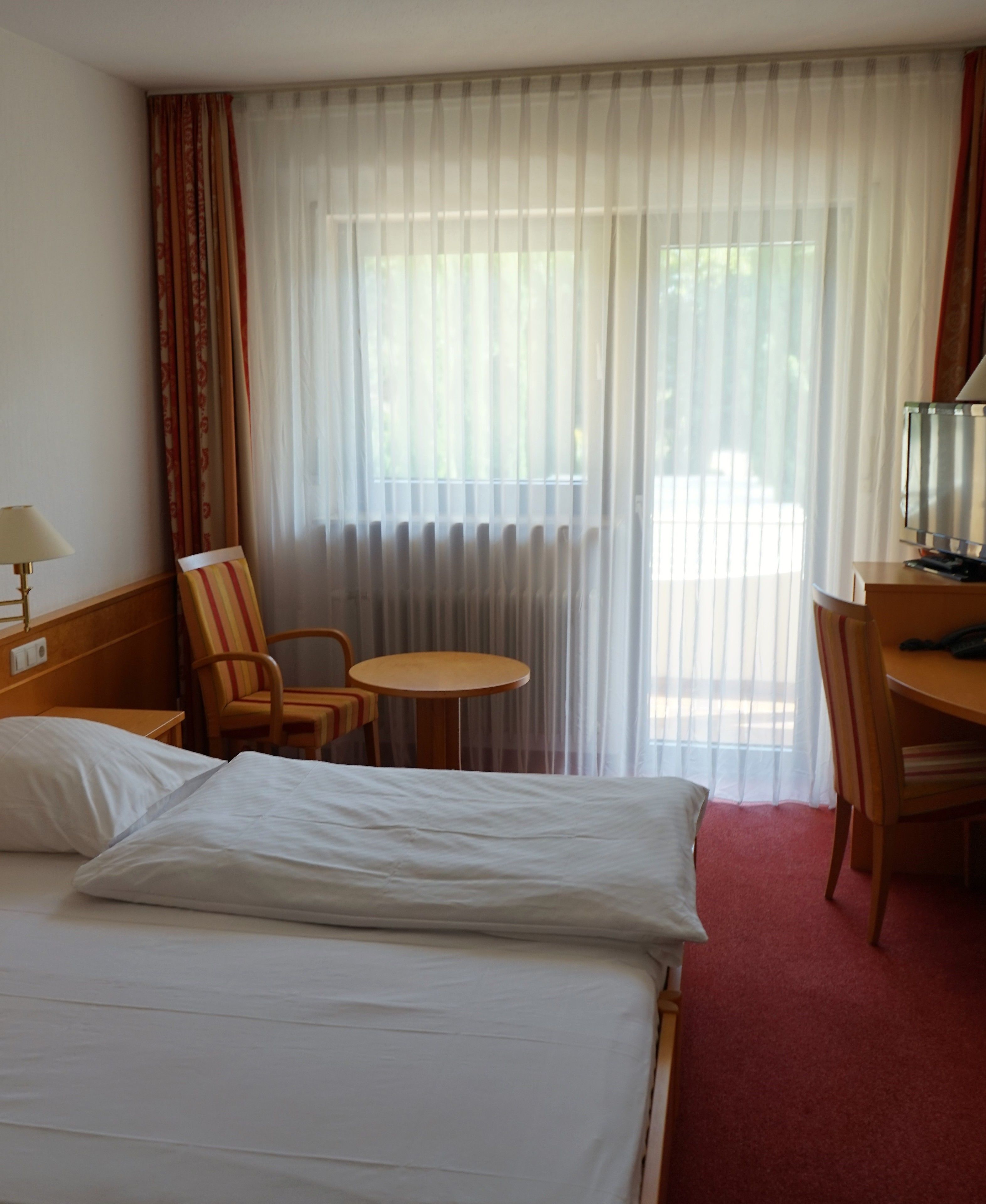 Une chambre d’hôtel avec un lit, un bureau, une chaise, une petite table et une fenêtre avec des rideaux.