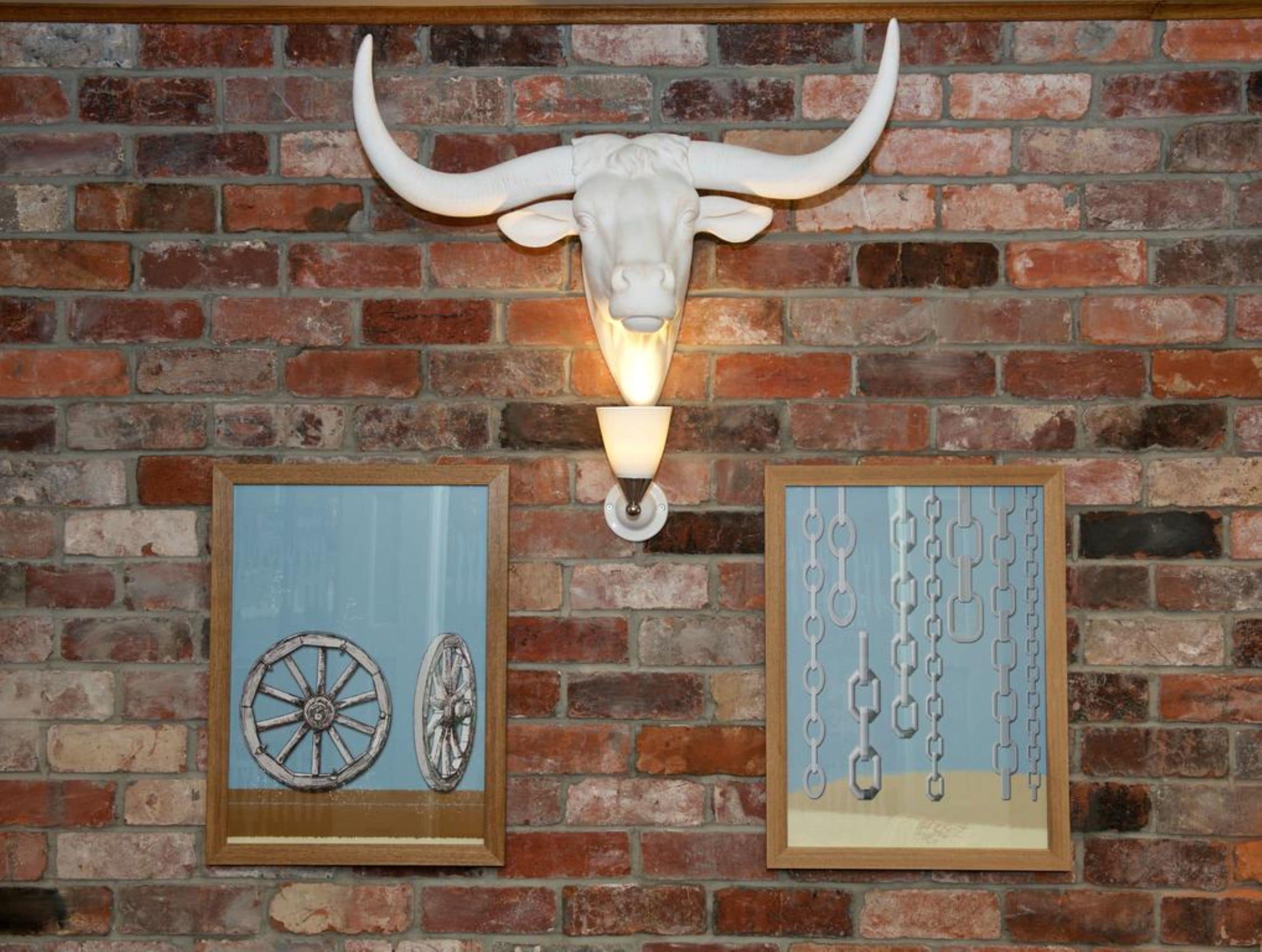 Een bakstenen muur met daaraan bevestigd een koehoofd en twee ingelijste kunstwerken eronder.