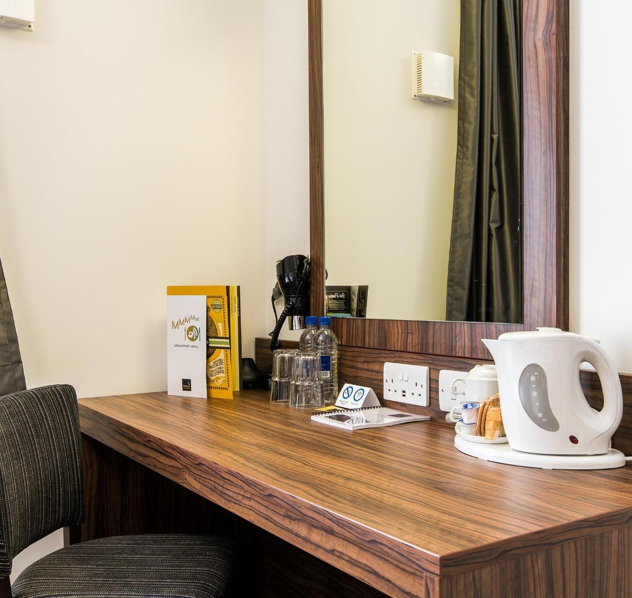 A hotel room with a wooden desk, a coffee maker, a kettle, a water bottle, a notepad, and a breakfast menu box.