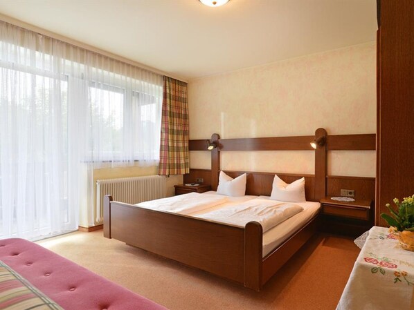 Een slaapkamer met een houten bed, een raam met dunne gordijnen, een radiator en een nachtkastje met een lamp.