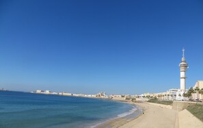Un paisaje urbano costero con un faro destacado, playa de arena y cielo azul despejado.