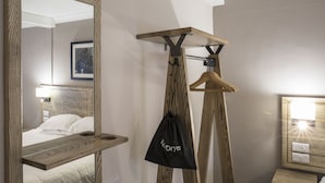 Une chambre d’hôtel avec un grand miroir, une tête de lit en bois, un porte-vêtements avec un sac noir, et une table de chevet avec une lampe.