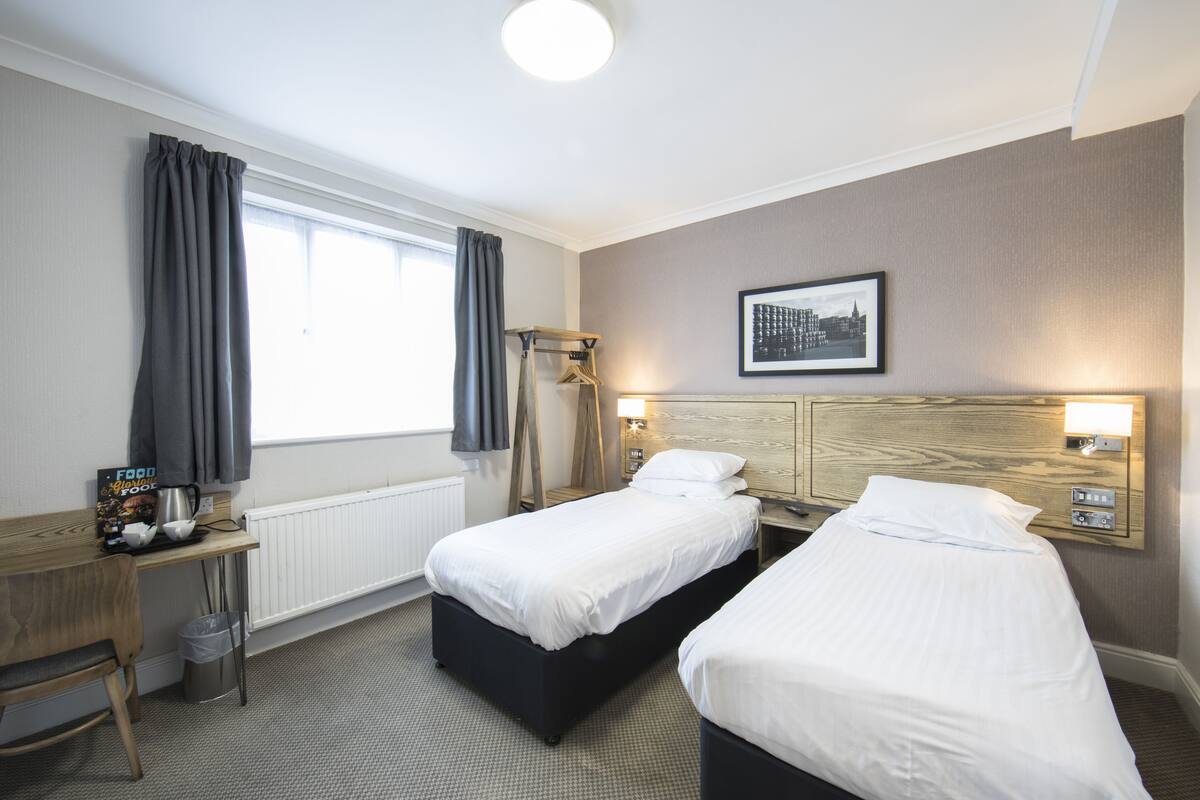 A hotel room with two single beds, a wooden desk, a chair, a radiator, and a framed picture on the wall.