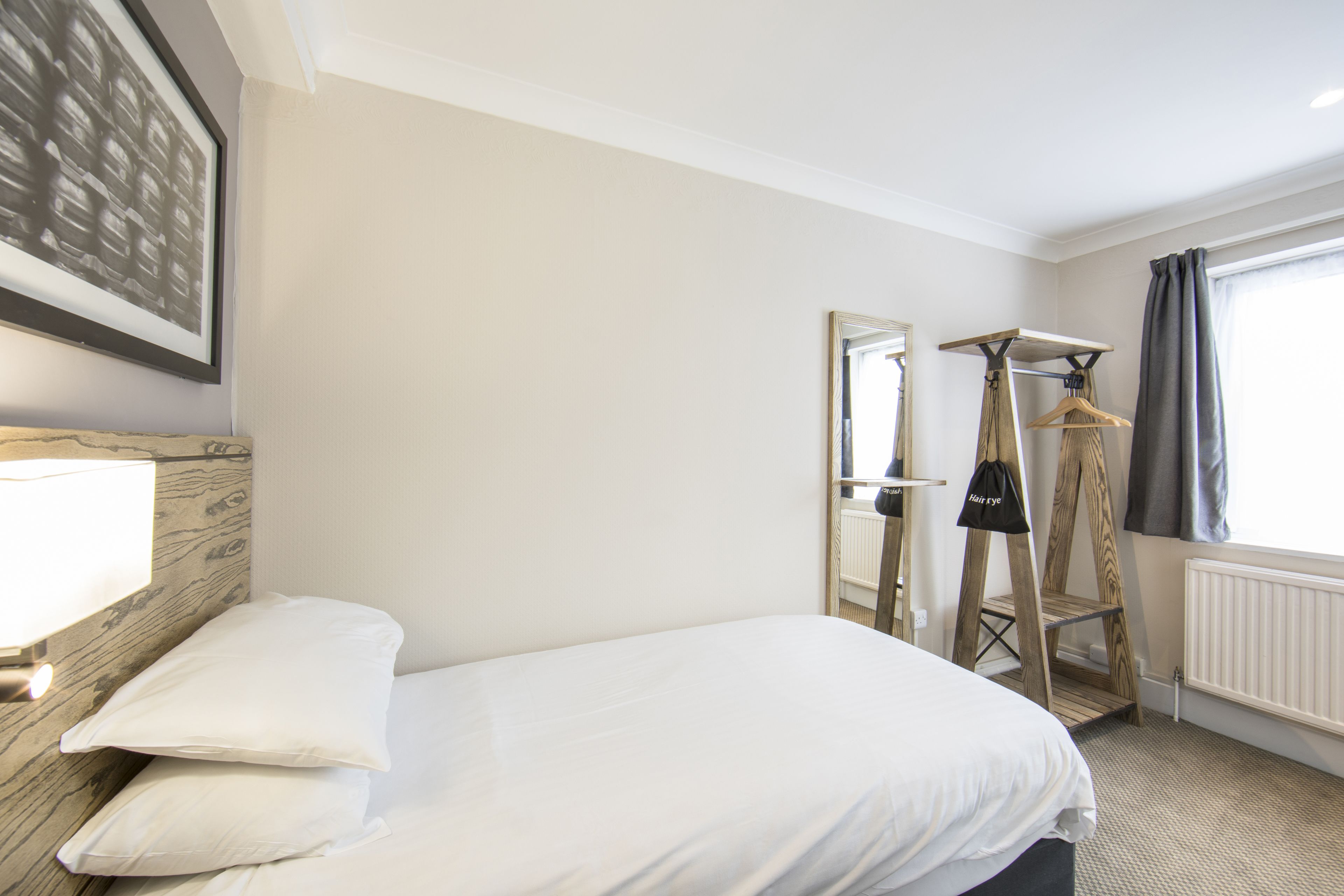 Habitación de hotel con cama, cabecero de madera, lámpara colgante, espejo, radiador y ventana con cortinas.