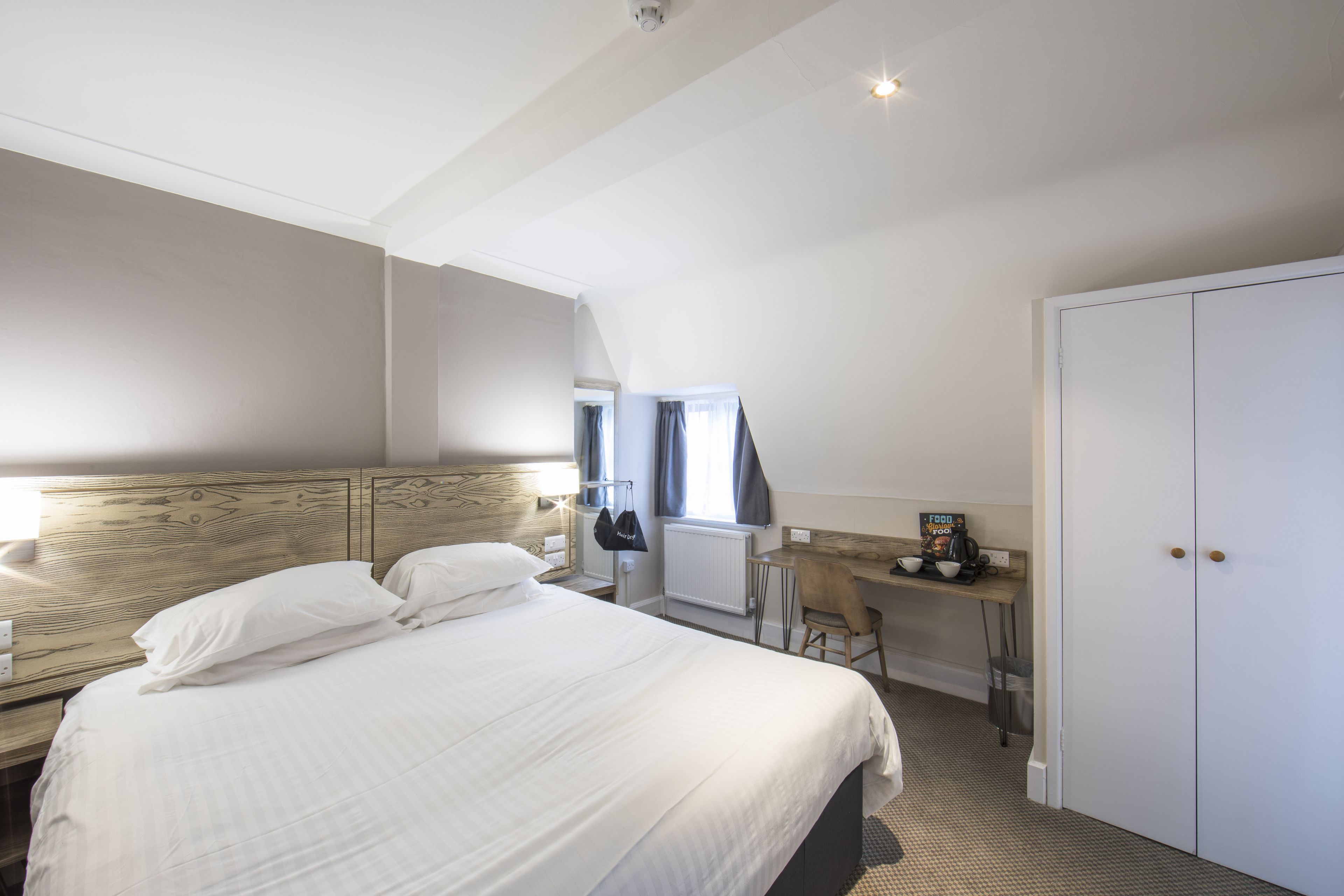 A hotel room with a large bed, a desk with a chair, a radiator, and a wardrobe.