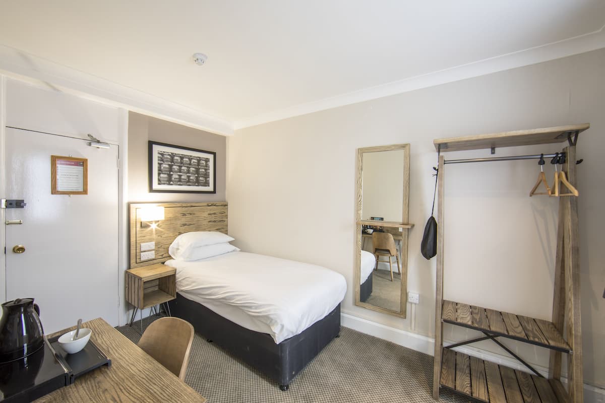 A hotel room with a bed, bedside table, chair, and a wooden shelf unit.