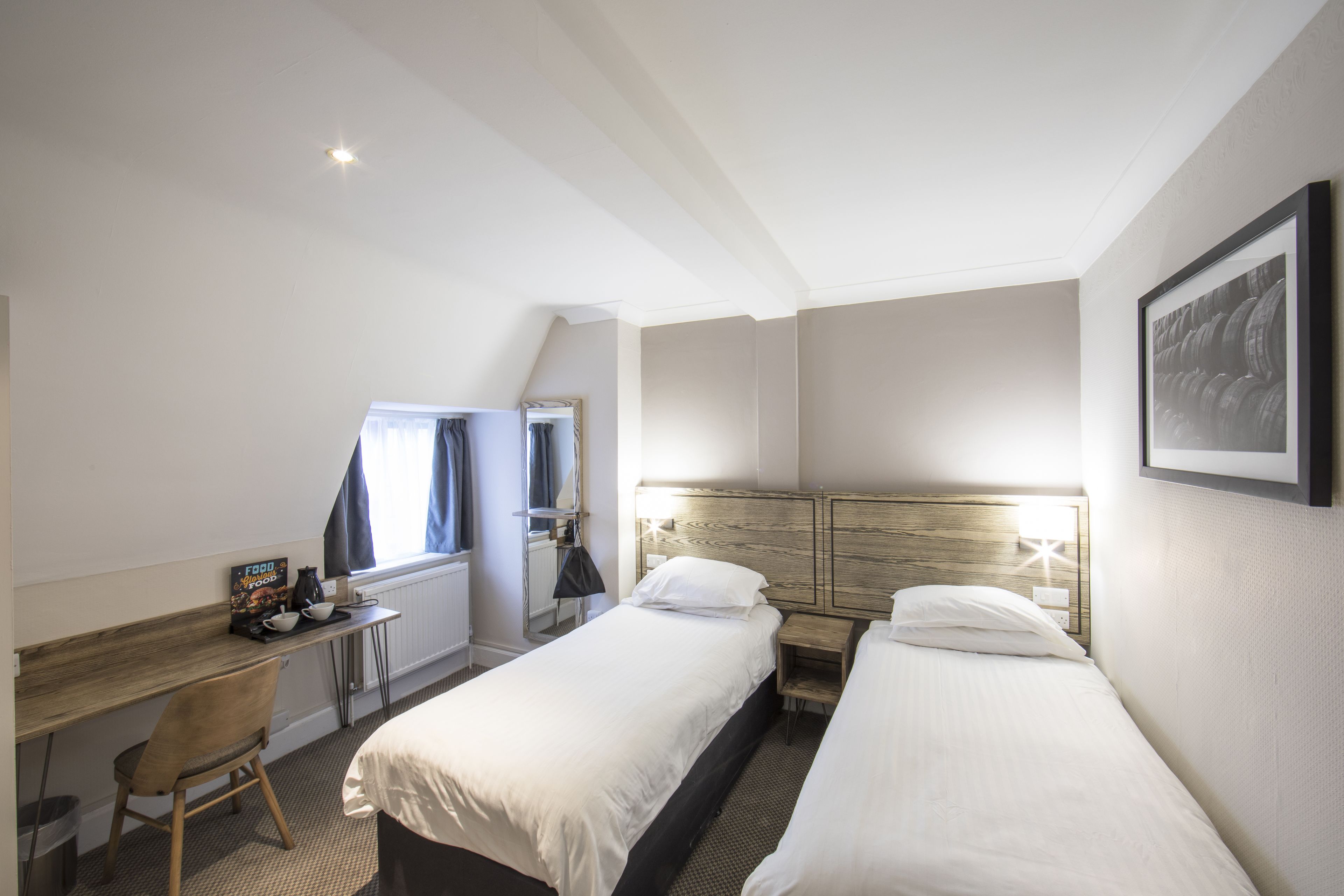 A hotel room with two single beds, a desk with a chair, a small table, and a radiator.