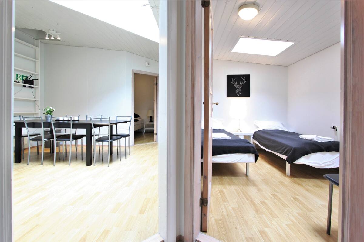 A room with a bed, bedside table, and a desk with chairs. There is a door leading to another room.