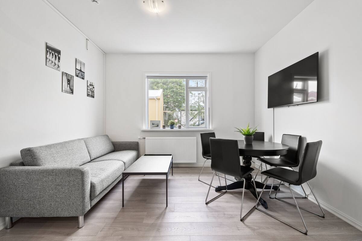 A modern living room with a grey sofa, a small table, and a dining area with chairs.