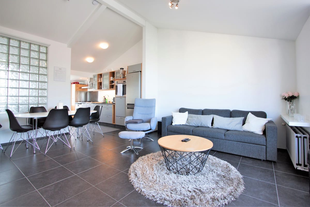 A modern living room with a grey sofa, round coffee table, and black chairs. A kitchen is visible in the background with a fridge and oven.