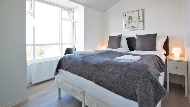 A bedroom with a large window, a bed with a grey blanket, a bedside table with a lamp, and a framed picture on the wall. - Odinn Reykjavik Odinsgata Apartments (Reykjavik)