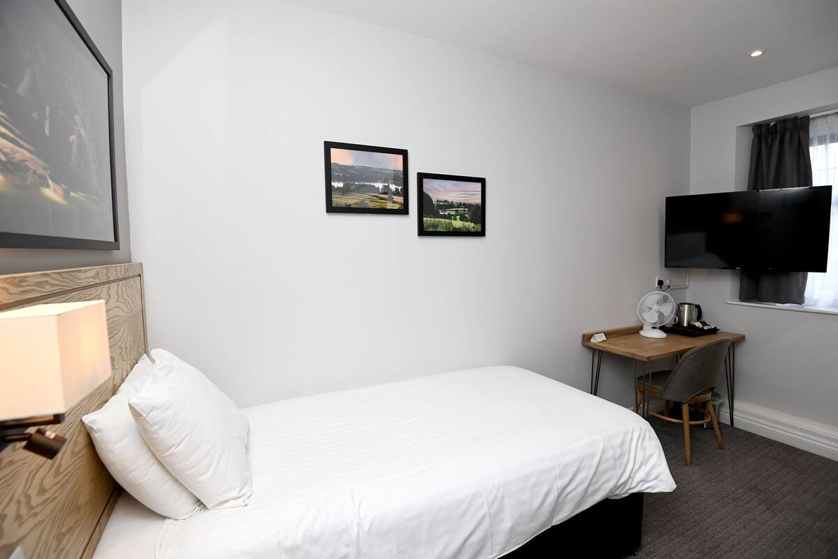 A hotel room with a bed, a desk with a chair, a television, and two framed pictures on the wall.