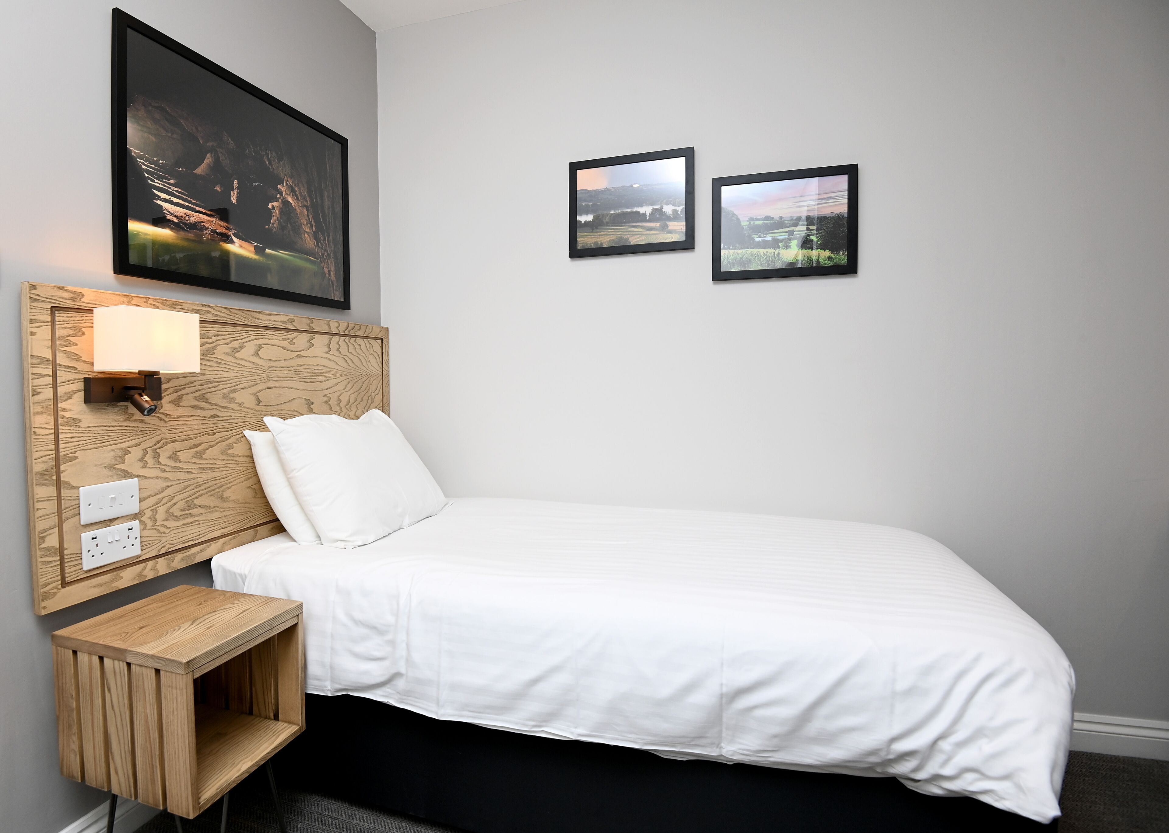 A neatly made bed with white bedding, a wooden headboard with a built-in light, and two framed pictures on the wall.