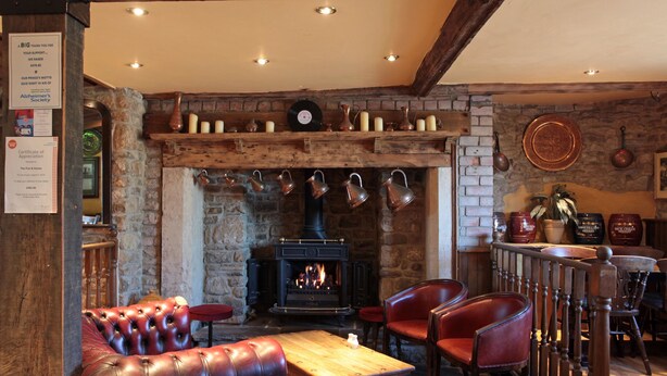 A cozy pub with a stone fireplace, leather seating, and a wooden bar area. - Fox & Goose, Barrow Gurney by Marston's Inns (Bristol)