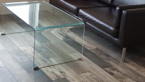 Una sala de estar con una mesa de centro de vidrio, un sofá de cuero marrón y vista a los coches estacionados afuera a través de una ventana.